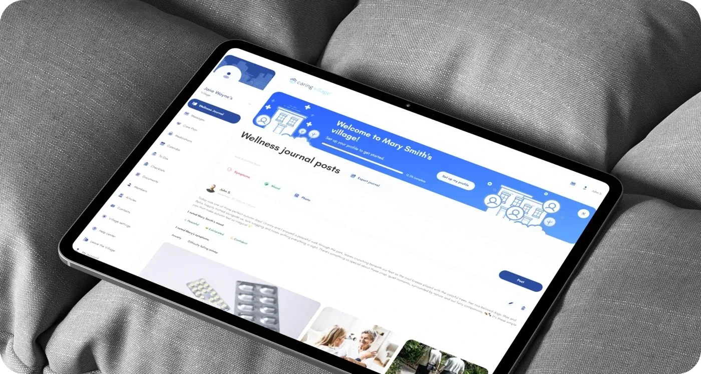 A tablet resting on a grey cushion showing the redesigned Caring Village web platform with the 'Wellness journal posts' feed and a 'Welcome to Mary Smith's village!' banner.